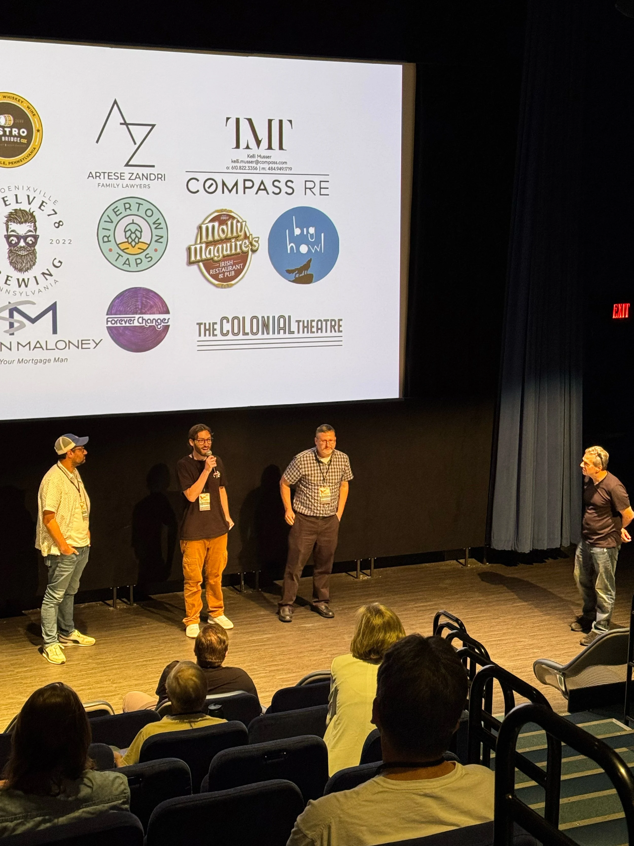 People standing on stage in front of an audience during a presentation in a theater, with a large screen displaying various logos.