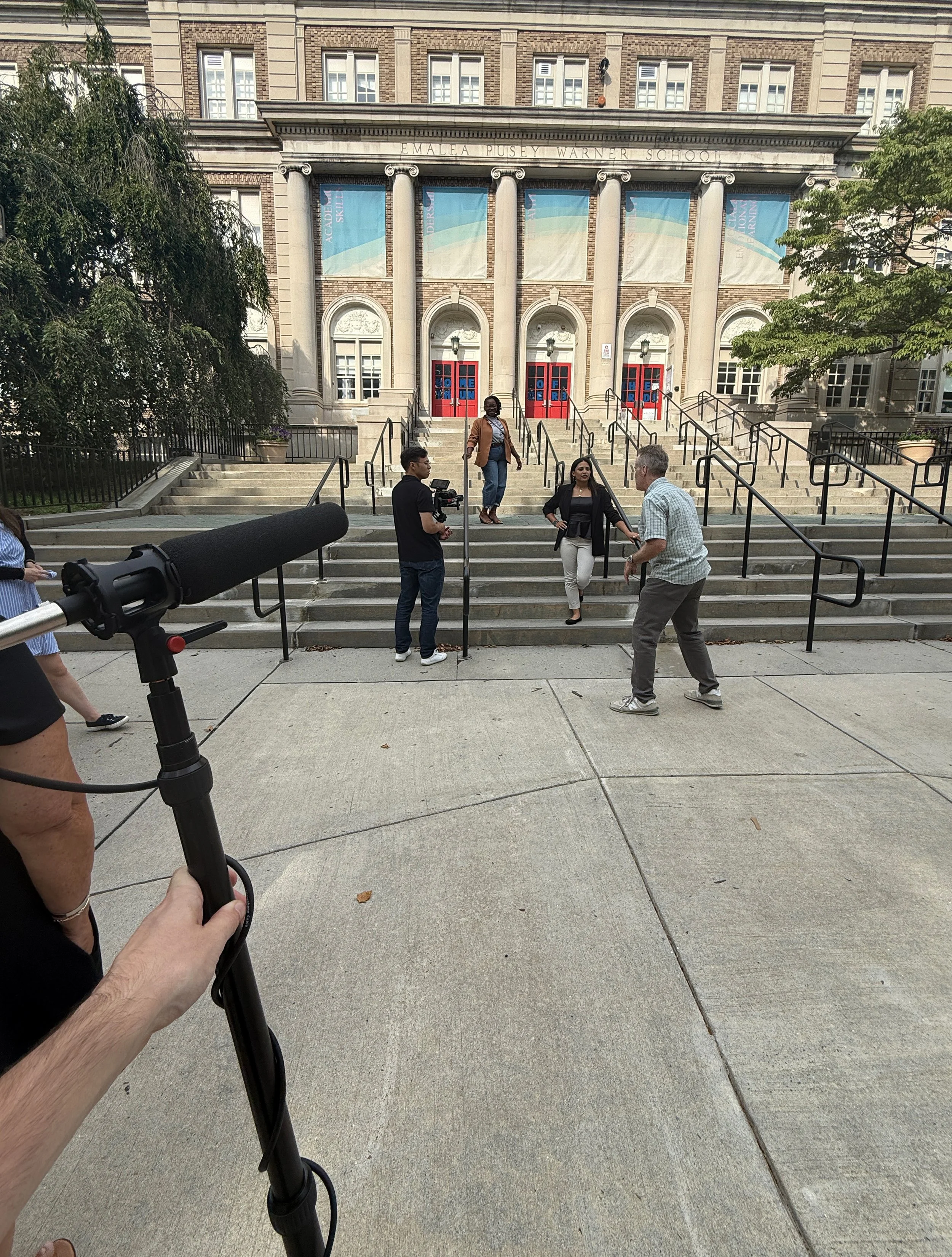 A building with a classical architectural style has stairs leading up to red doors and columns. Several people are standing outside, including a woman with a blazer and a man with a camera. There is a large microphone and boom pole in the foreground.