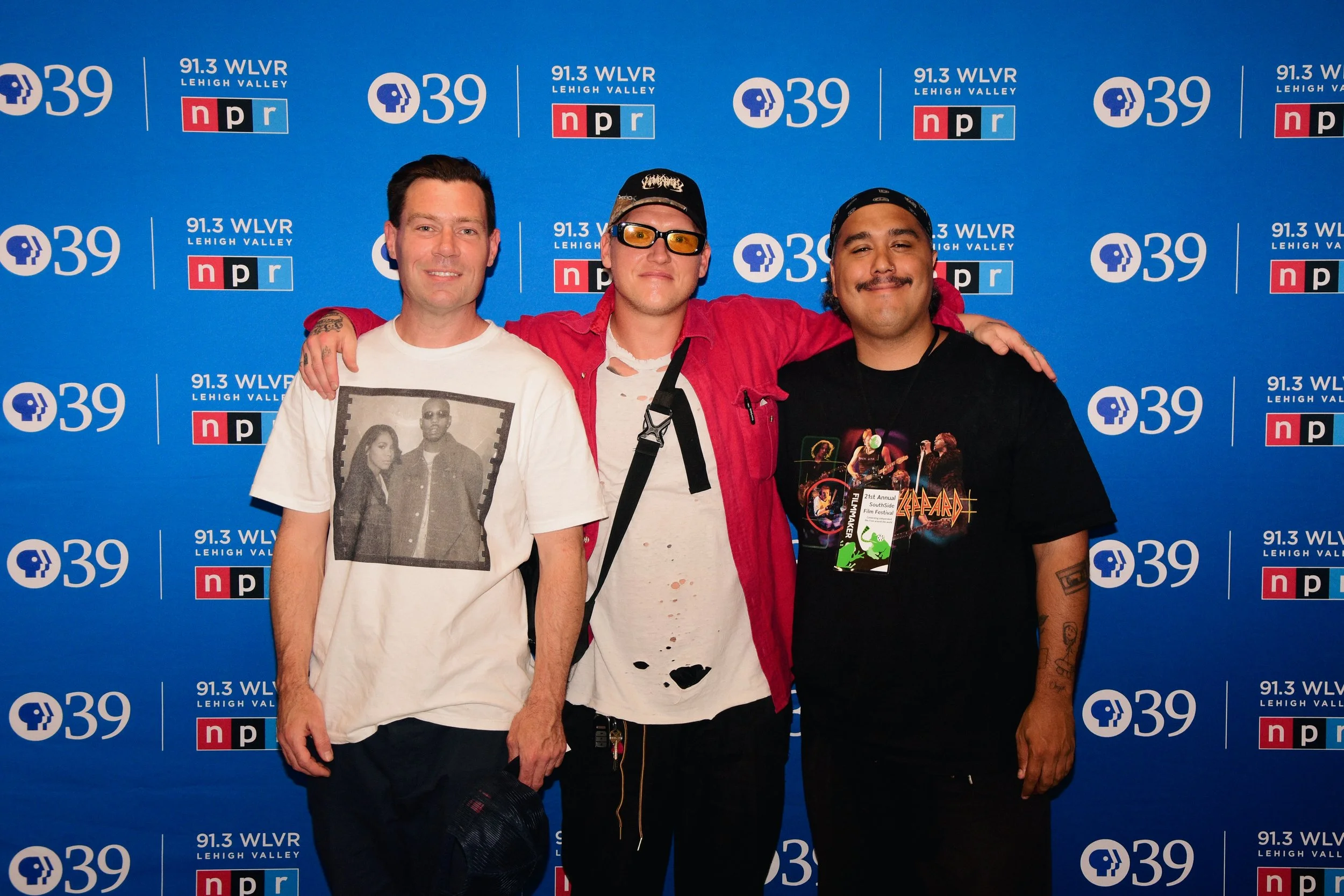 Three men standing arm-in-arm in front of a blue backdrop with radio station logos. All three are smiling; the man in the middle wears glasses and a red shirt, while the other two men wear graphic t-shirts.