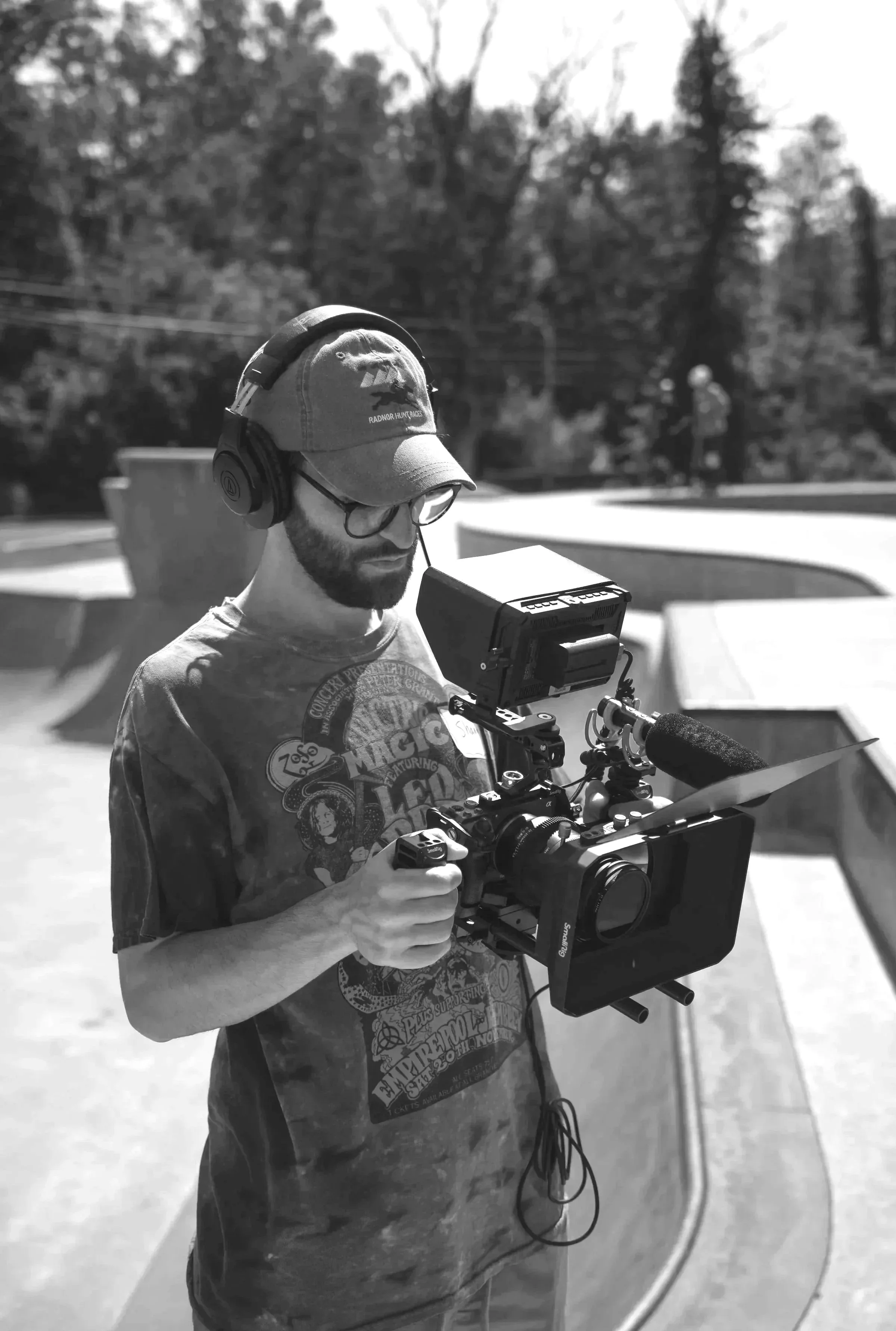 A man with glasses and a baseball cap operating a professional camera in an outdoor skate park, with trees and a person in the background.