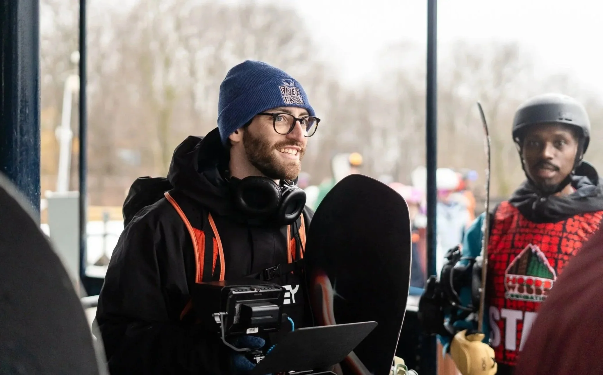 Two men in outdoor winter gear, one with a blue beanie and glasses and the other with a helmet, standing at a counter with a snowboard and coaching or preparing for snowboarding in a winter outdoor setting.