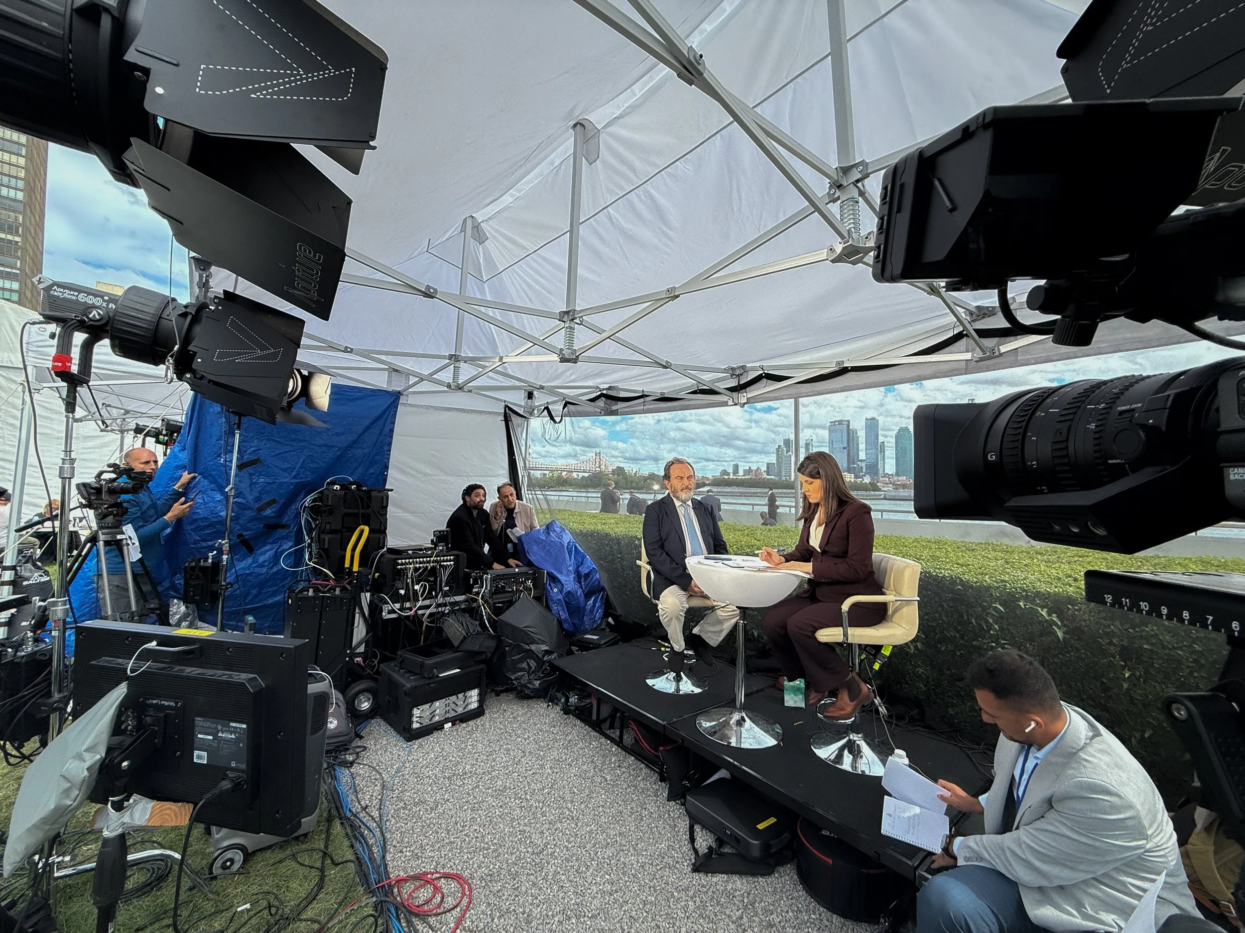 Filming crew conducting interviews outdoors under a white canopy with city buildings and skyline in the background.