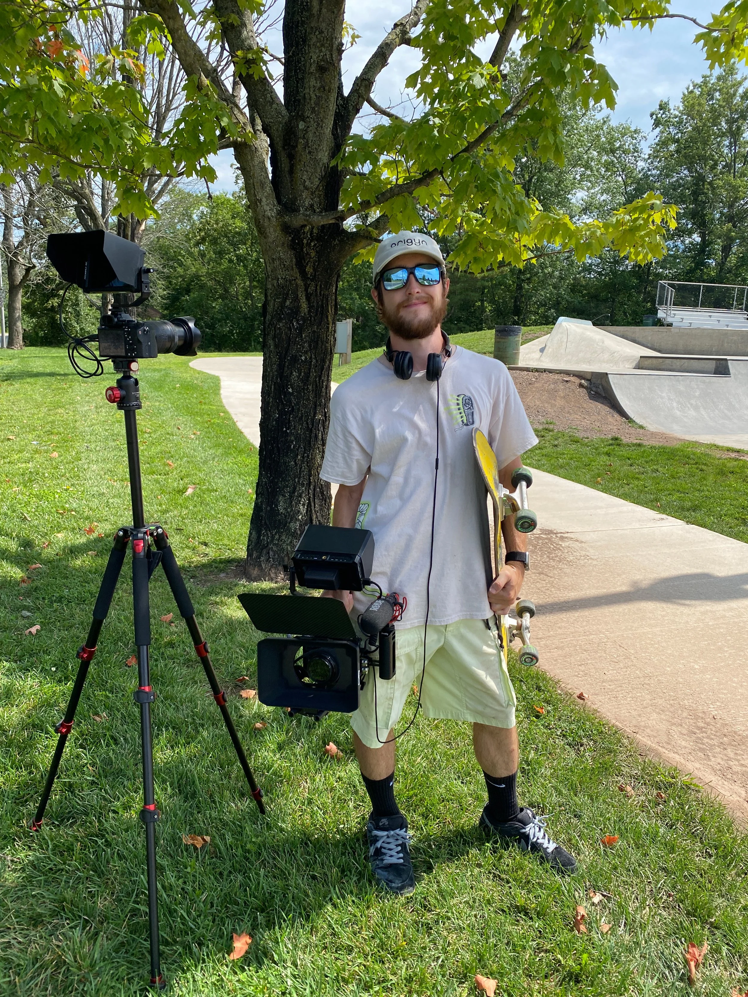 A young man with a beard, wearing sunglasses standing outdoors on a grassy area next to a tree, with a camera in hand. There is video recording equipment set up on a tripod nearby, and skateboarding ramps are visible in the background on a sunny day.