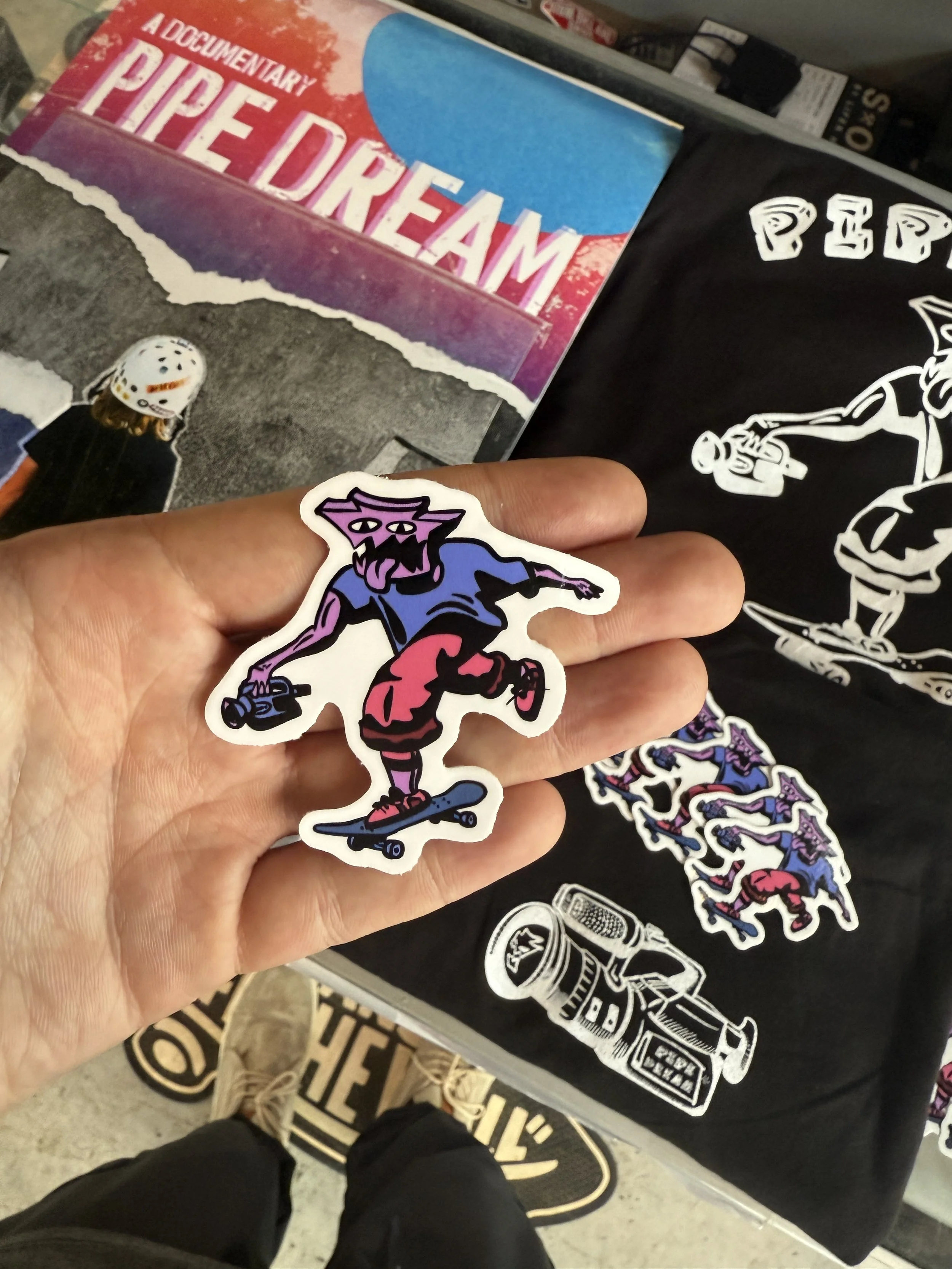 Close-up of a hand holding a purple and black sticker of a stylized cartoon character skateboarding. In the background are stickers and posters, including one with the words 'Pipe Dream' and another with a skeleton holding a spray paint can.
