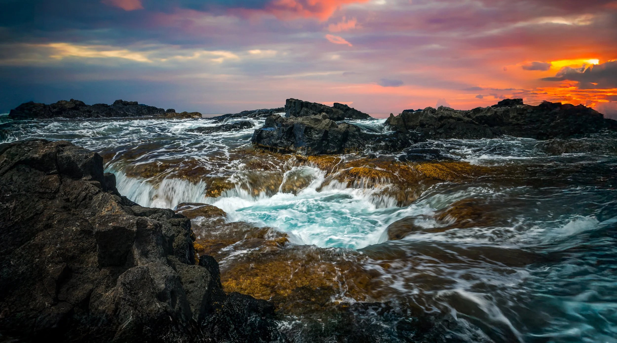 A rocky shoreline with ocean waves crashing against the rocks at sunset, with a colorful sky featuring orange, pink, and purple clouds.