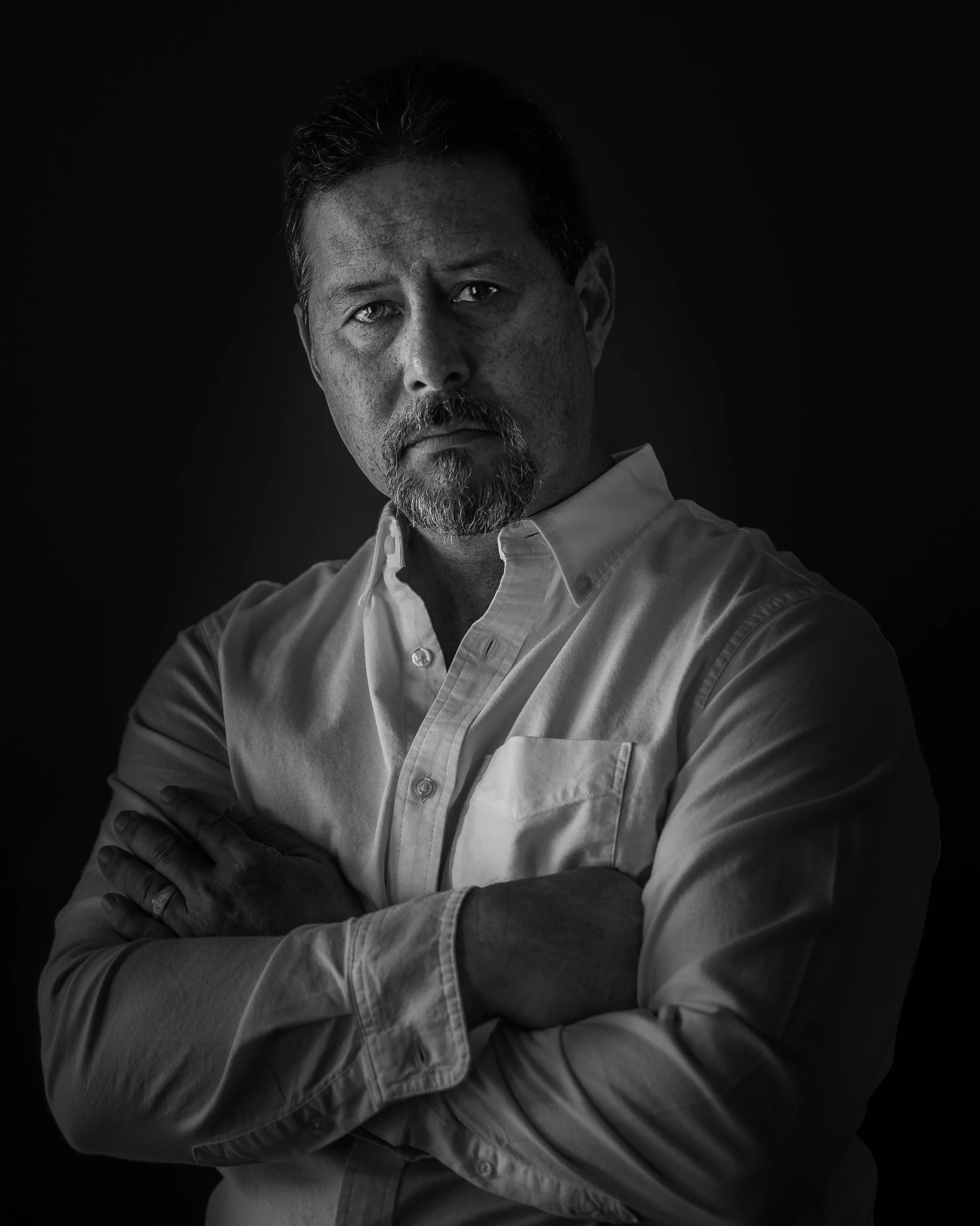 Black and white portrait of a man with a beard and mustache, wearing a collared shirt, with arms crossed, looking directly at the camera against a dark background.