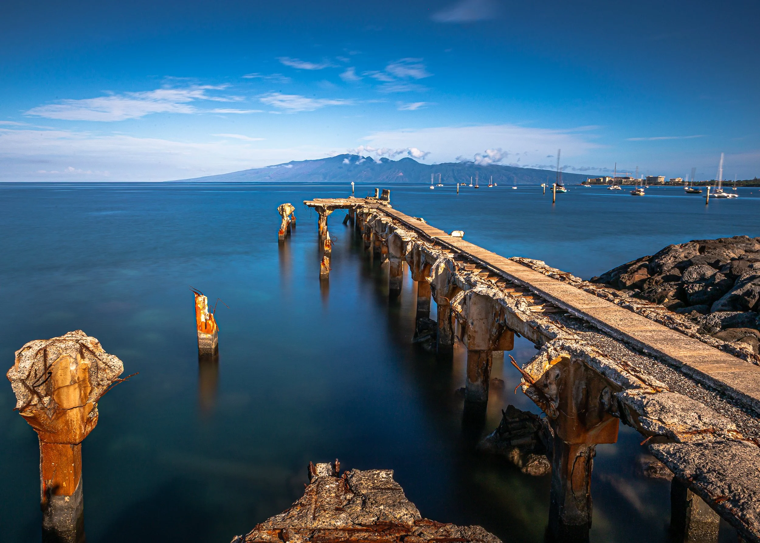 A long, broken wooden pier extending into calm water with boats anchored nearby, mountains in the distance, and a partly cloudy blue sky.