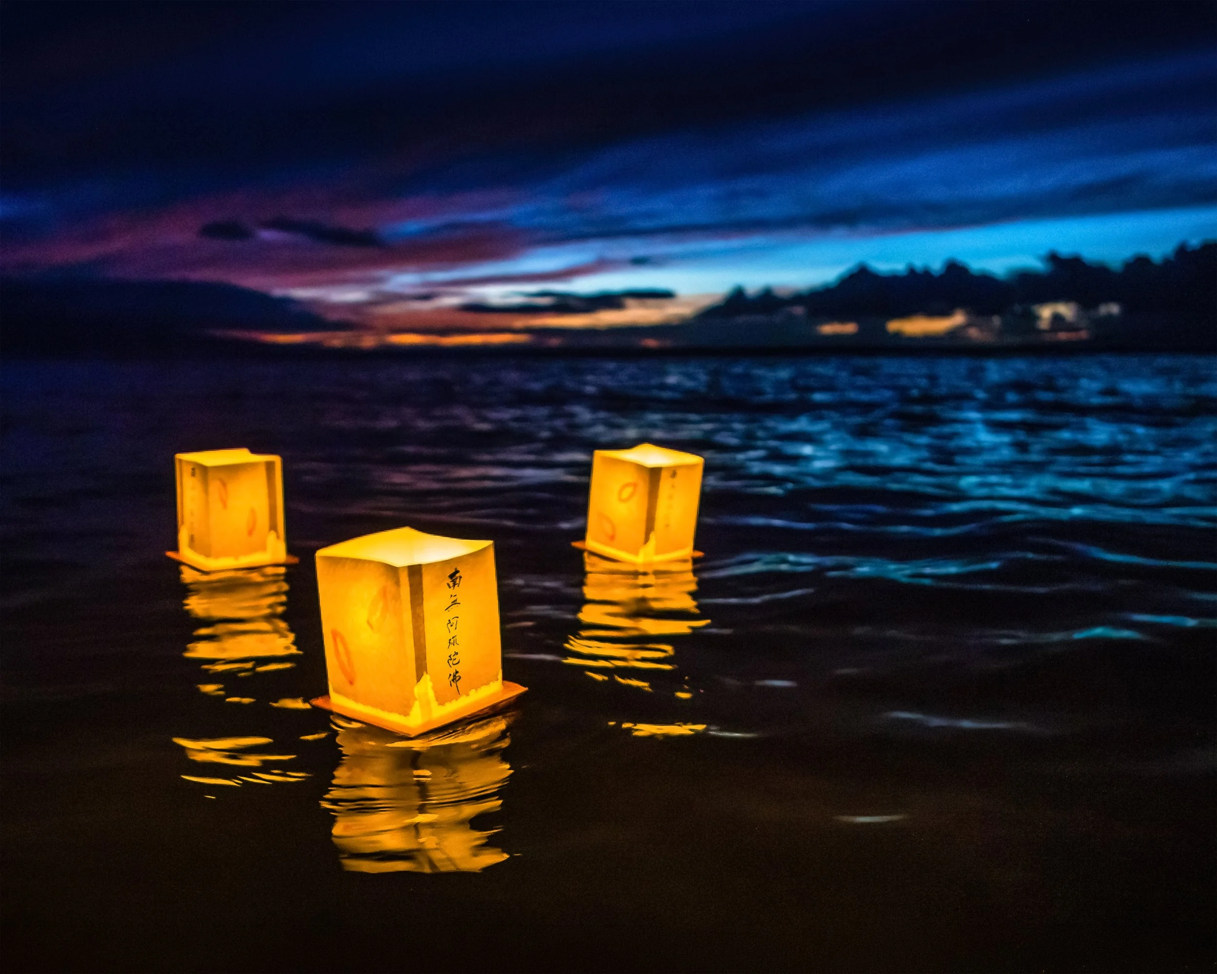 Three floating paper lanterns illuminated with yellow light on a body of water at dusk, with a colorful sunset and dark clouds in the sky in the background.