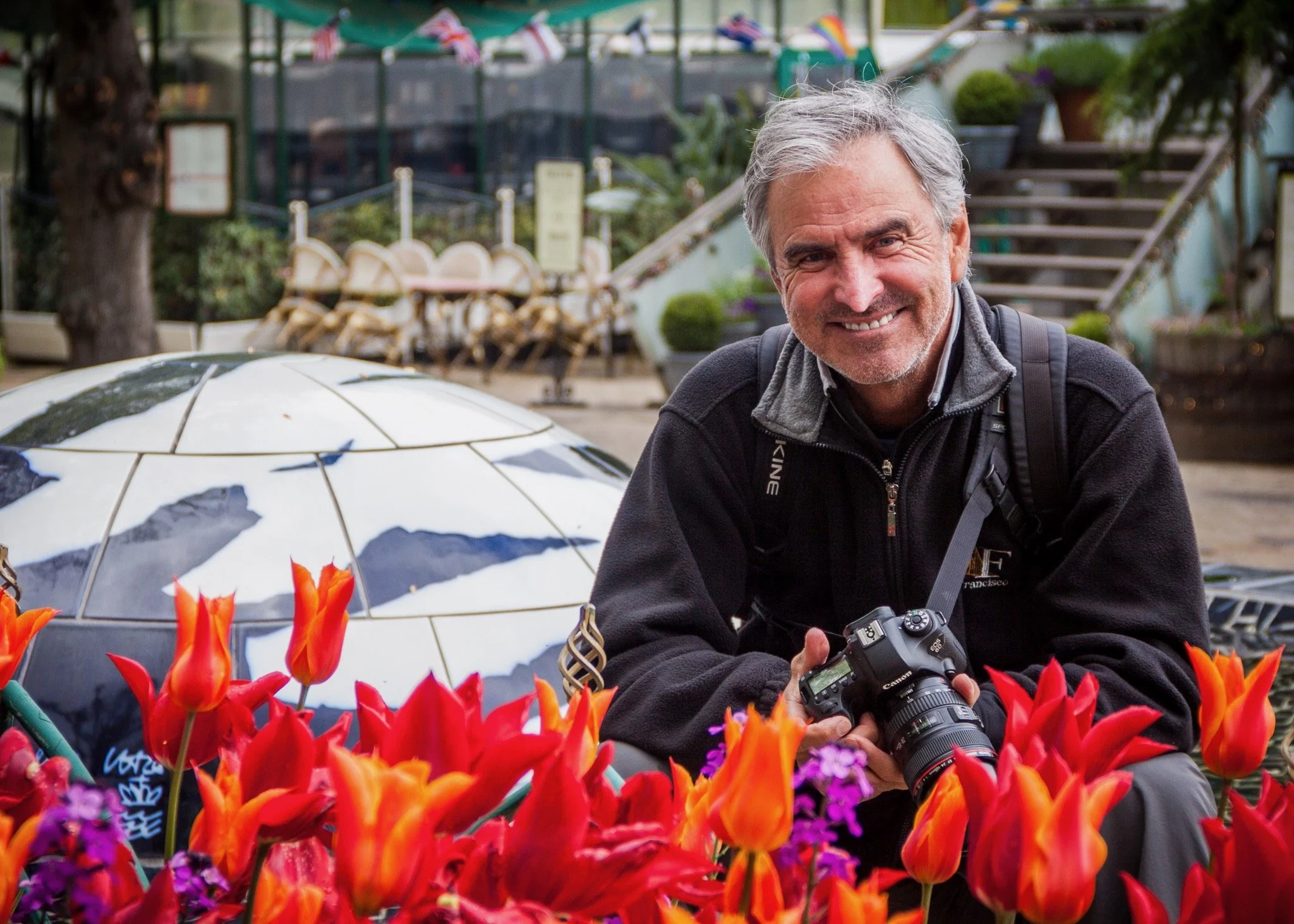 Smiling middle-aged man with gray hair and a beard holding a camera, surrounded by colorful red, purple, and orange flowers in a garden setting.