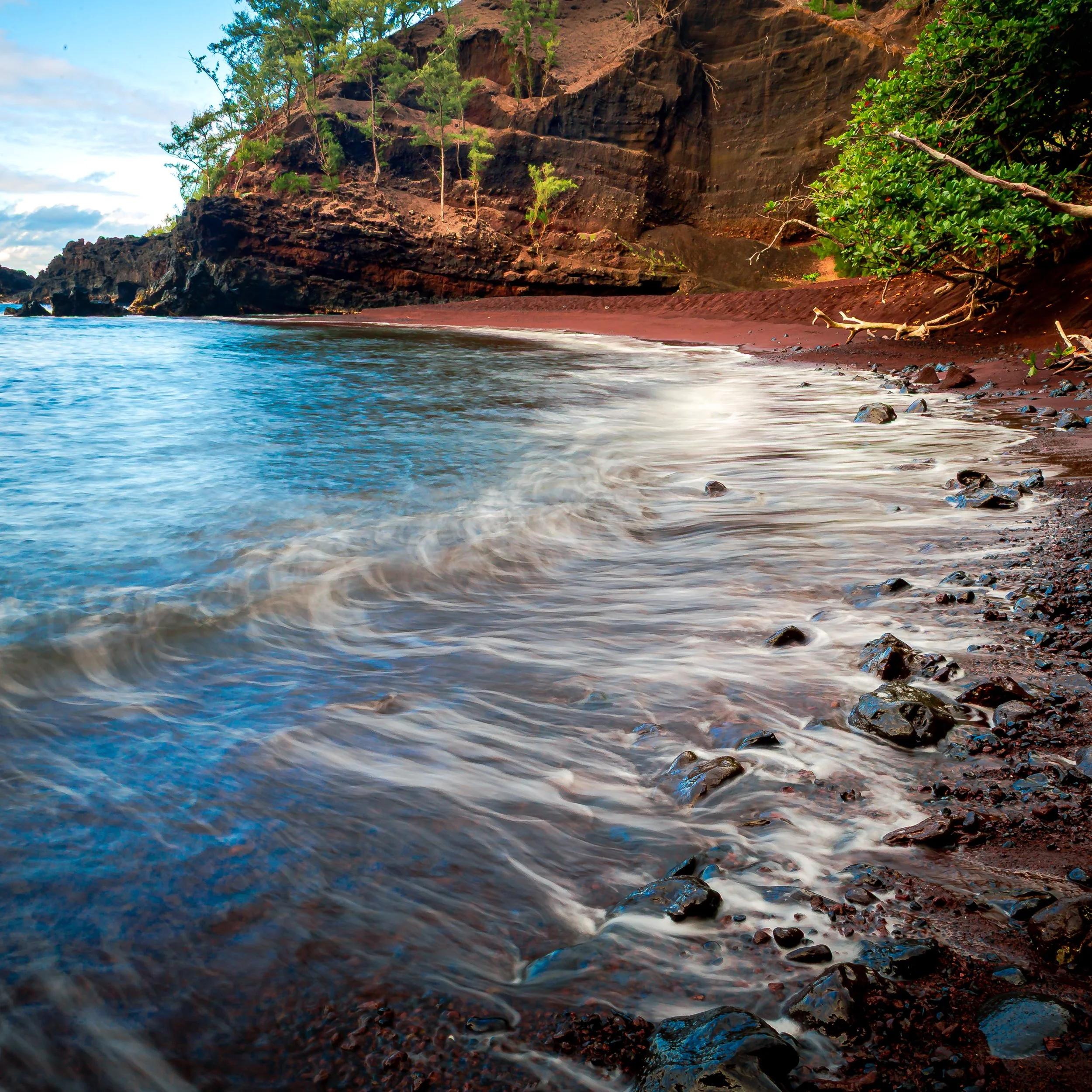 View of a rocky shoreline with red sand, trees, and a large cliff in the background under a partly cloudy sky.
