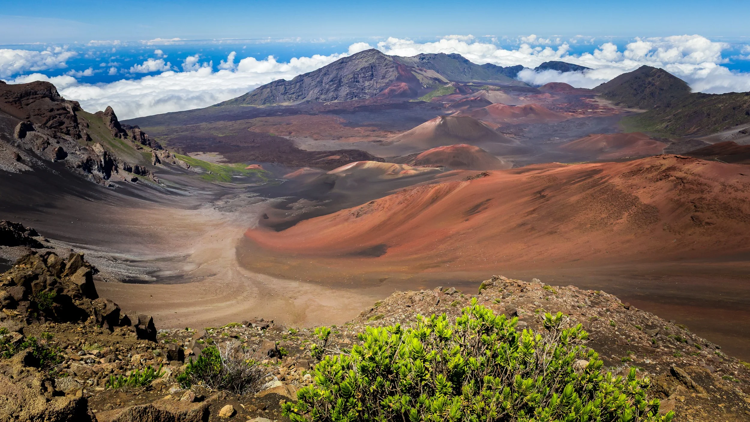 A volcanic landscape with reddish-brown slopes, volcanic craters, and dark lava fields, surrounded by clouds and distant mountains.