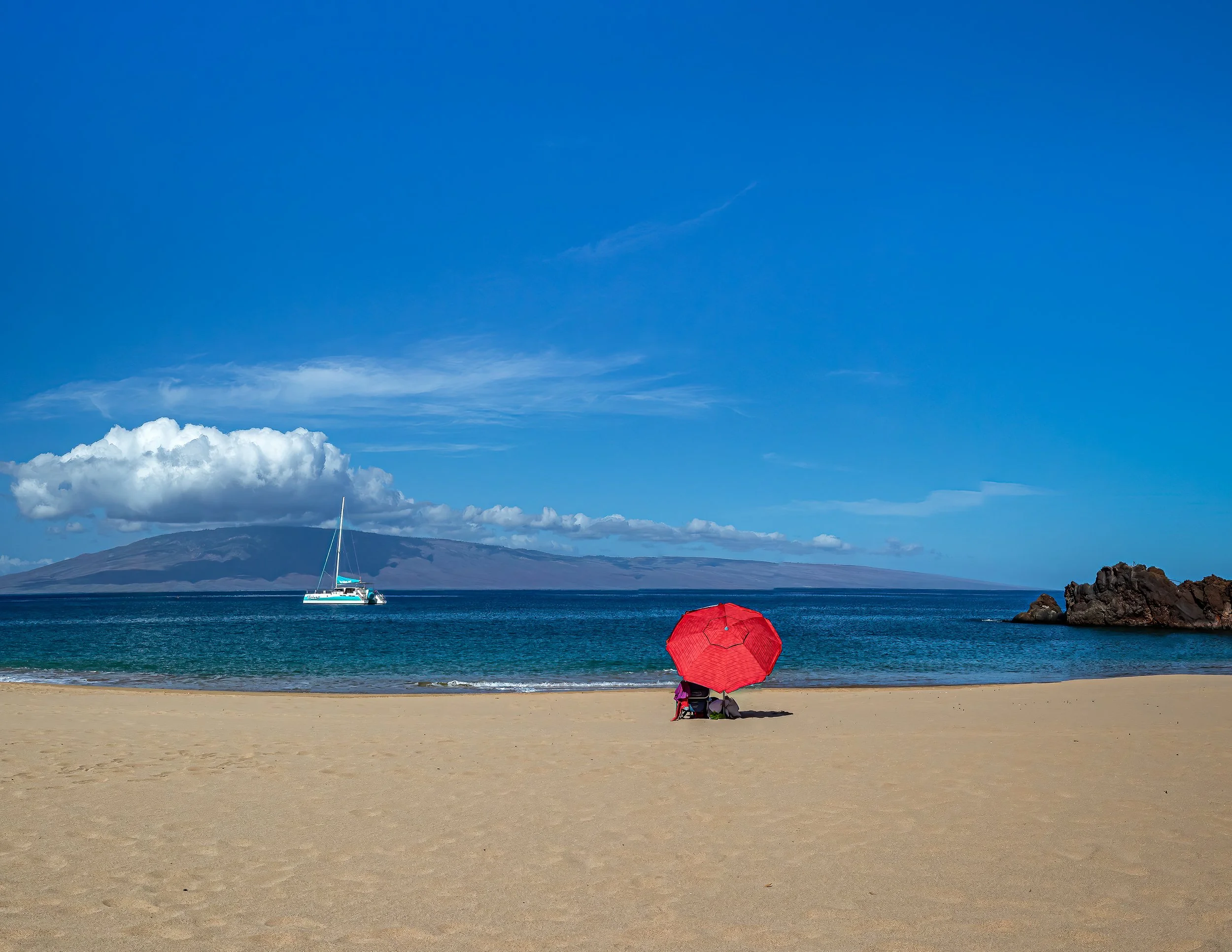 A person sitting on the sandy beach under a pink umbrella, facing the ocean, with mountains and a sailboat in the distance under a bright blue sky with some clouds.