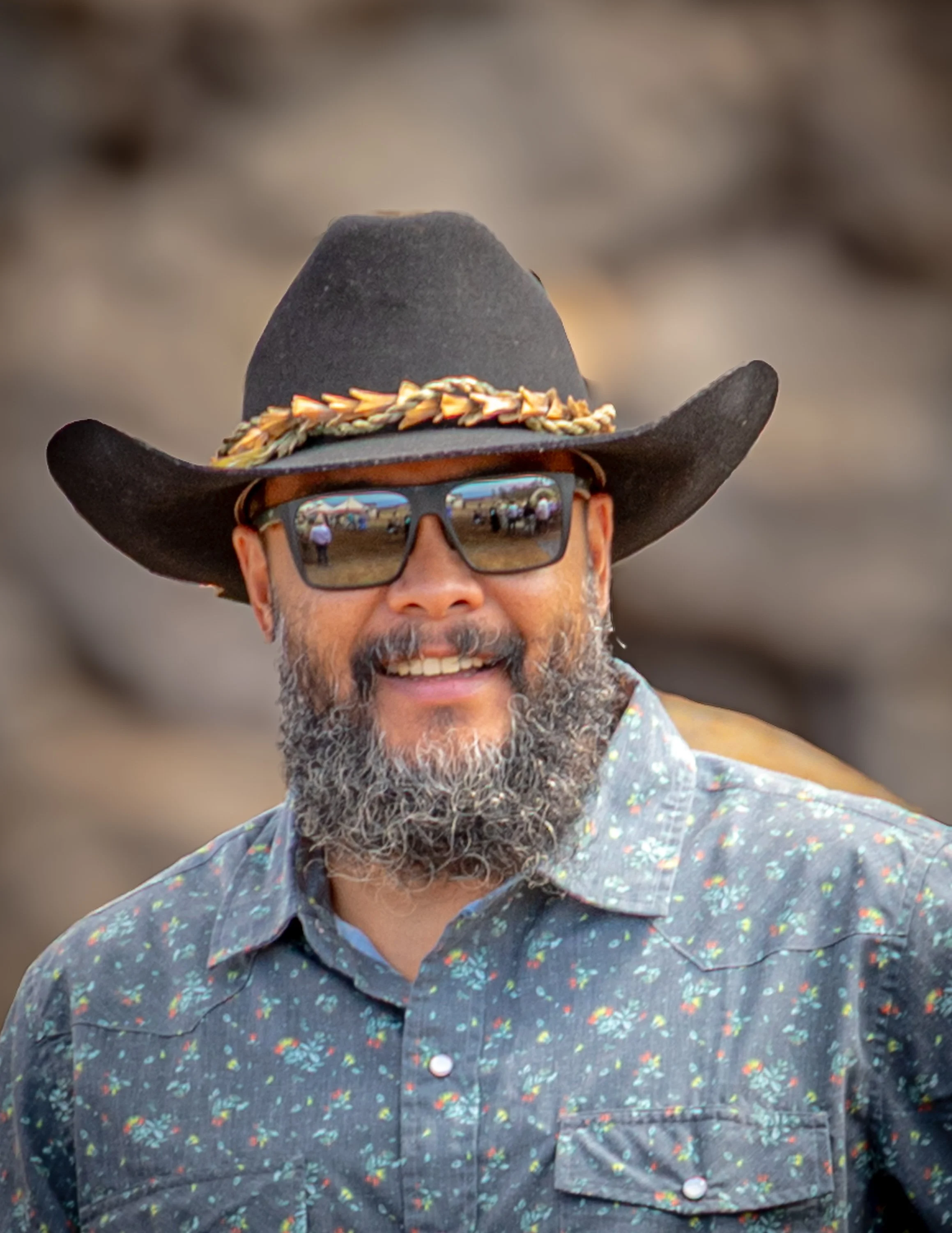 A man wearing a large black hat with a decorative band, sunglasses with reflections of people and tents, a beard, and a floral button-up shirt.