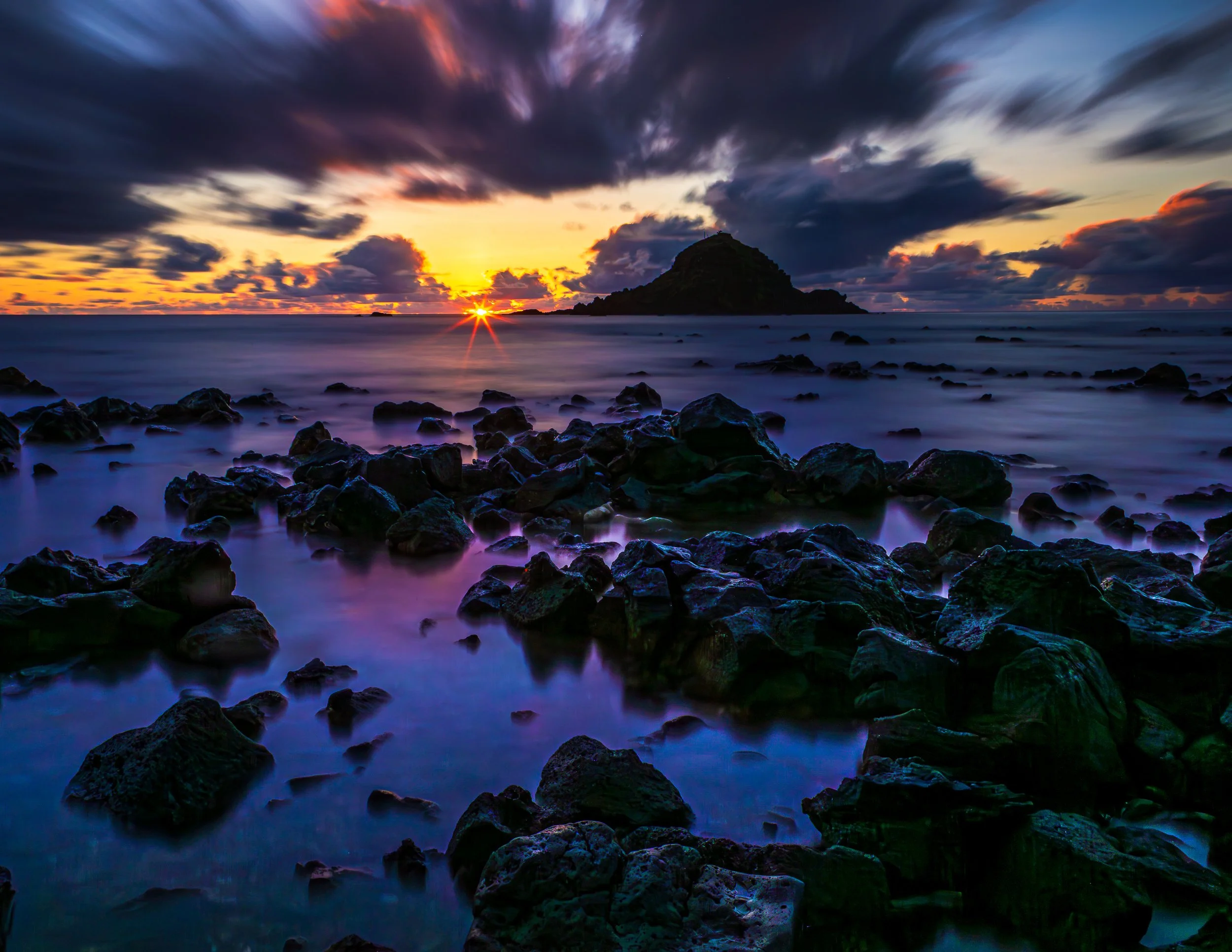 Sunset over a rocky coastline with a small island and dramatic clouds in the sky.