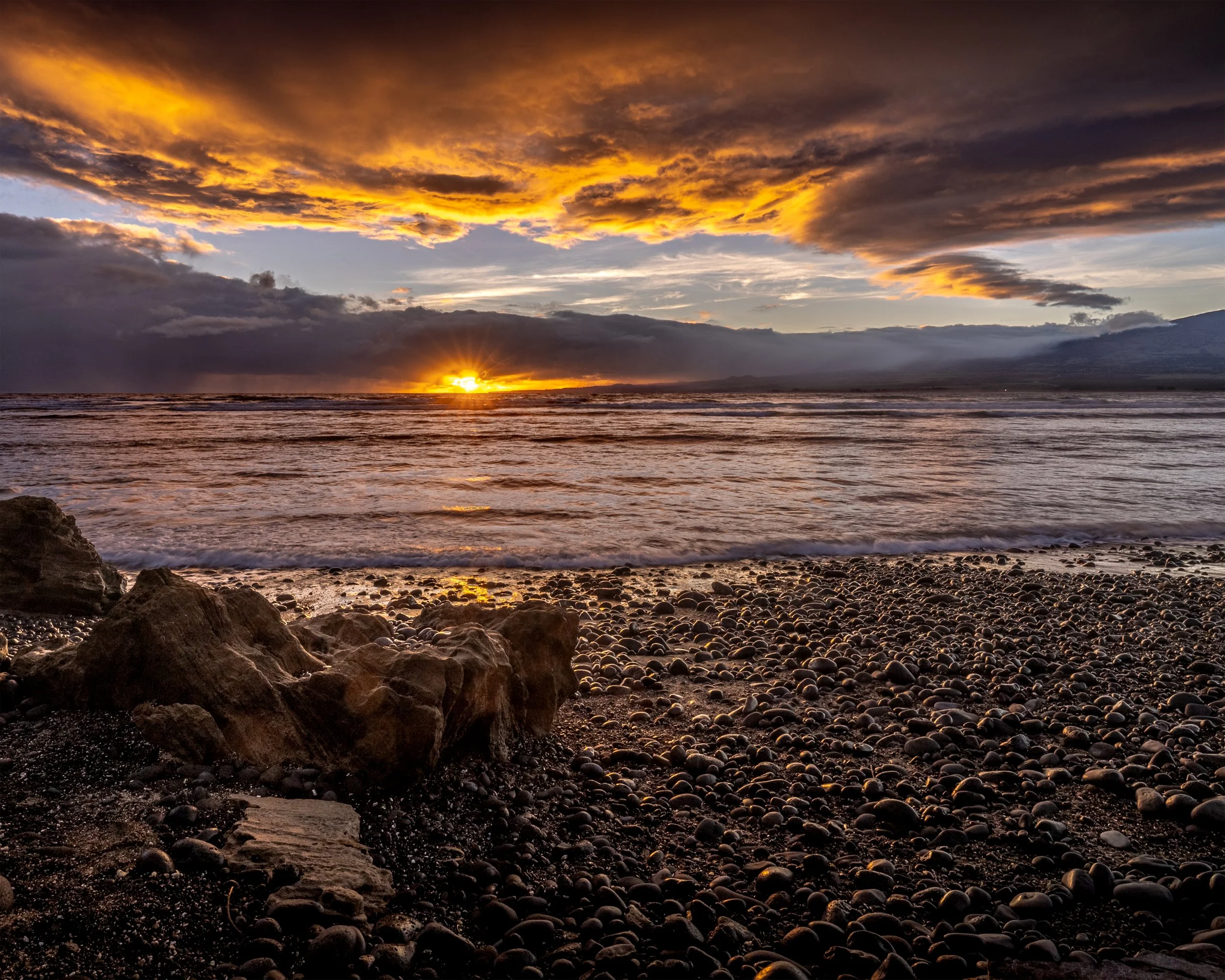 A scenic beach at sunset with dark clouds in the sky, displaying vibrant shades of orange and yellow near the horizon. Rocks and pebbles are seen along the shoreline, with the ocean waves gently coming in, and distant mountains are visible under the 