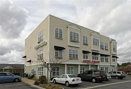 A three-story beige building with multiple windows, parking lot in front with several cars, and a sign that reads 'NIMMO EYECARE'.