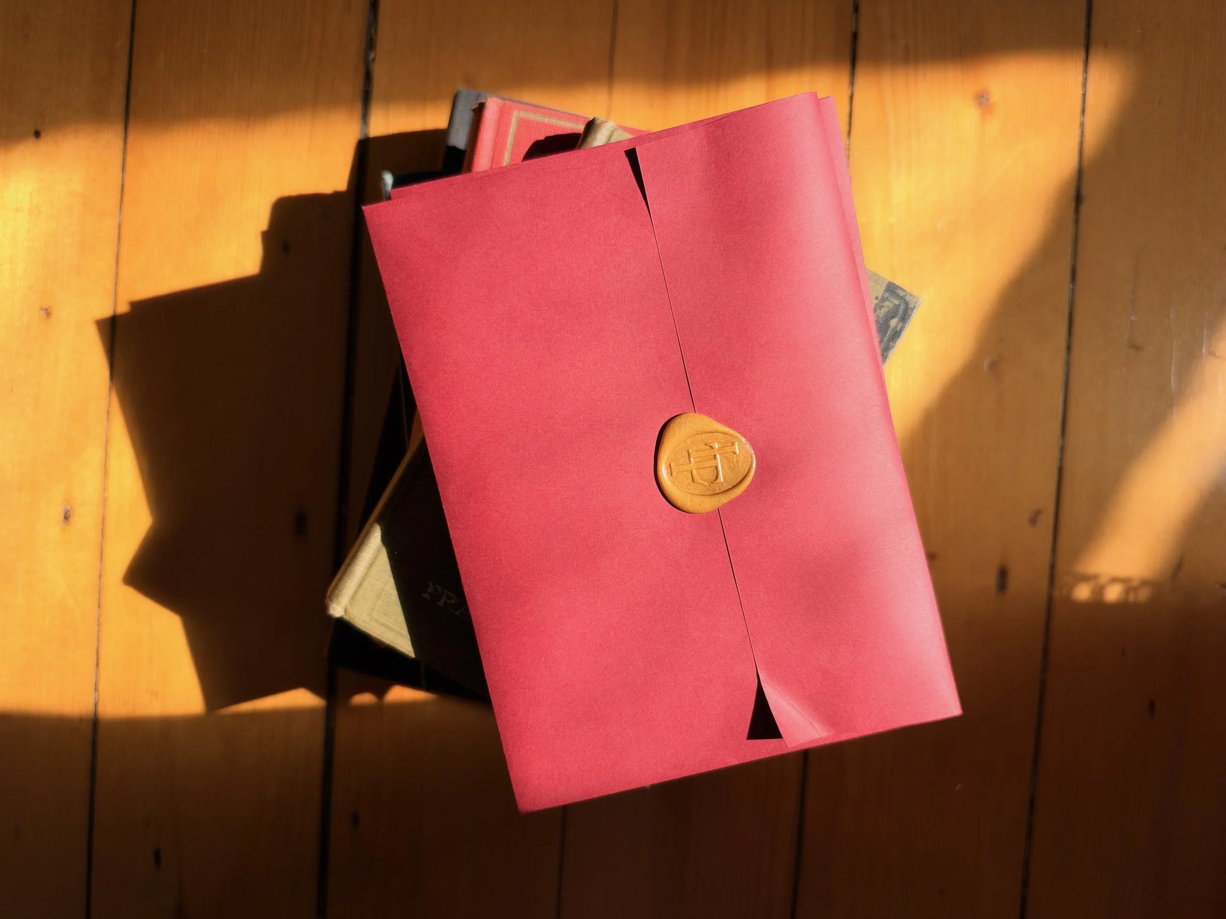 A red envelope with a gold seal on top, placed on top of a closed book, on a wooden floor with sunlight casting shadows.