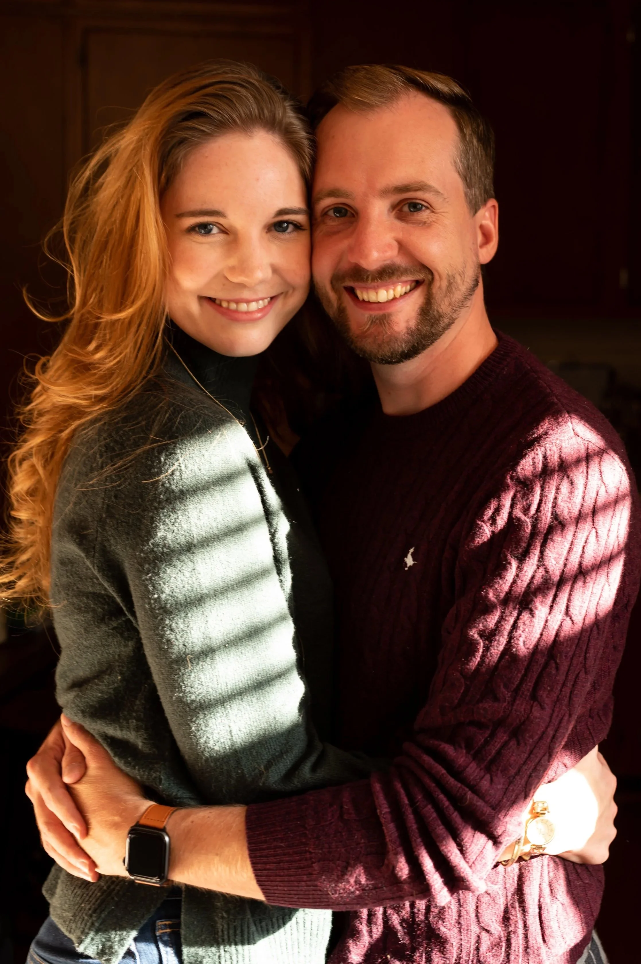 A man and a woman are hugging and smiling at the camera. The woman has long, wavy red hair and is wearing a gray and black sweater. The man has short brown hair, a beard, and is wearing a maroon sweater and a gold watch.