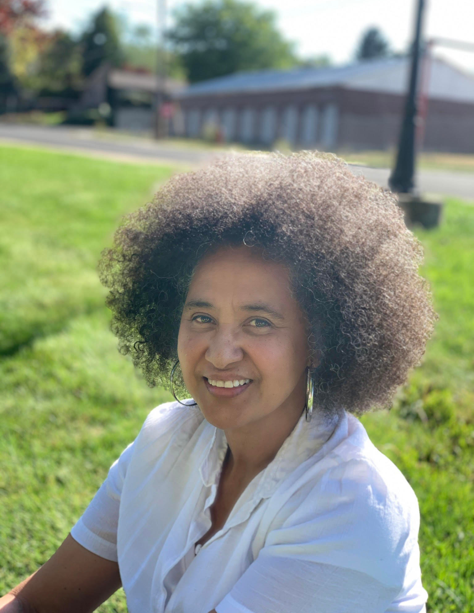 A woman with curly hair smiling outdoors on a sunny day, wearing a white shirt, with a building, trees, and a lamp post in the background.