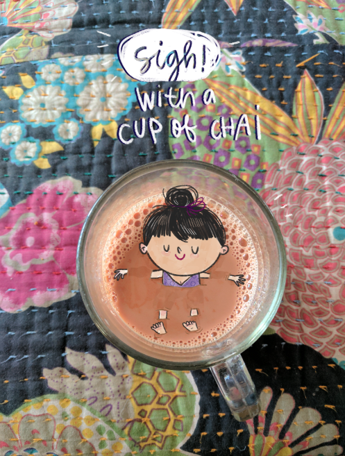 A glass of chai placed on a colorful quilt with floral patterns. The surface of the chai has an illustration of a girl with black hair tied in a bun, smiling with her arms and legs out. Above the glass, there is a speech bubble with the text 'sigh! with a cup of chai' written inside.