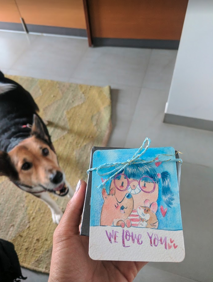 A hand holding a small gift box decorated with a drawing of a girl with blue hair, glasses, and a striped shirt, hugging two mice, with hearts and the words 'We Love You' written on it. A dog is looking up in the background, standing on a yellow-green rug near a kitchen floor.