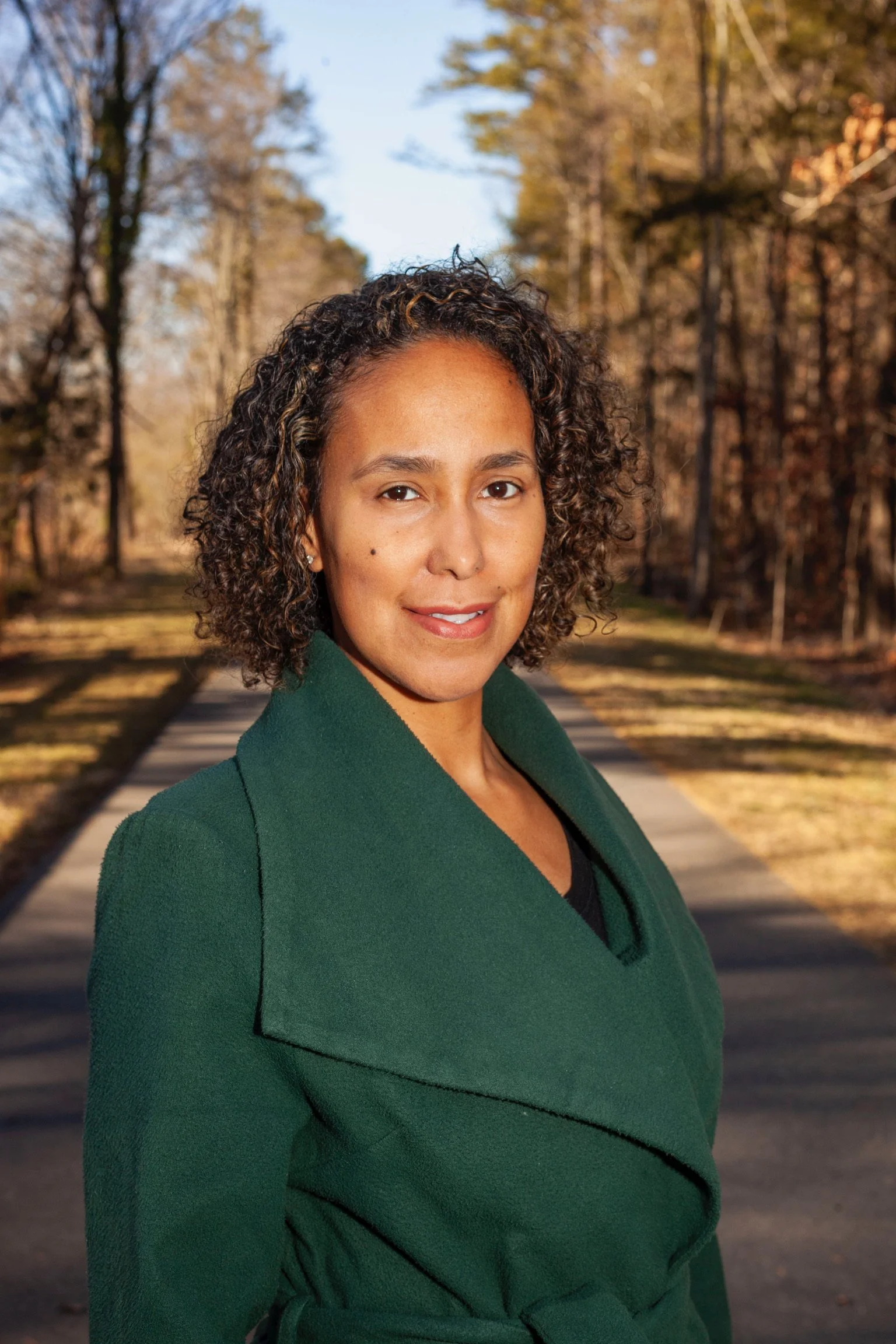 Sarah Peralta. www.manosdesarah.com. Charlotte, NC. A woman with curly hair wearing a green coat standing outdoors on a path surrounded by trees with autumn leaves.