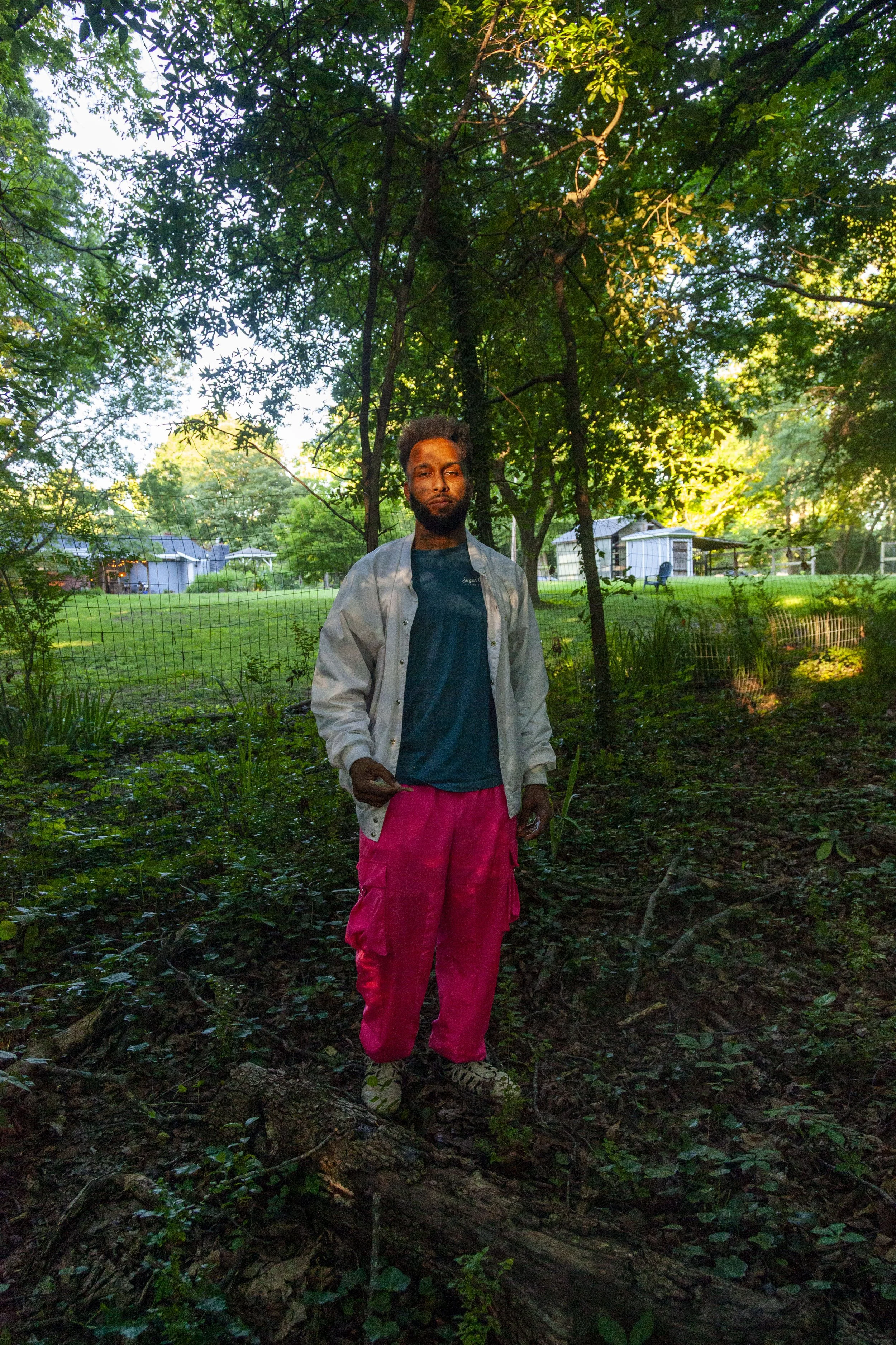 Logan Ellis. Charlotte, NC. A man with a beard standing outdoors in a wooded area during daytime. He is wearing a gray jacket over a dark shirt and bright pink pants. The background includes trees, a house, and a backyard with chairs and fencing.