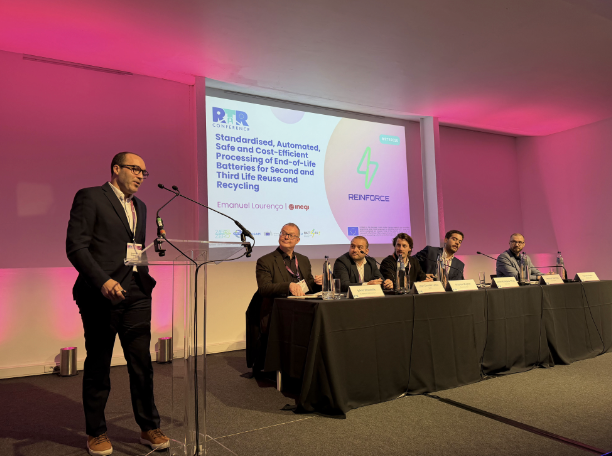 A man standing at a podium speaking at a conference, with five men seated at a table on stage. A presentation slide behind them displays the title 'Standardised, Automated, and Cost-Efficient Space and End-of-Life Batteries for Second and Third Life Reuse and Recycling.'