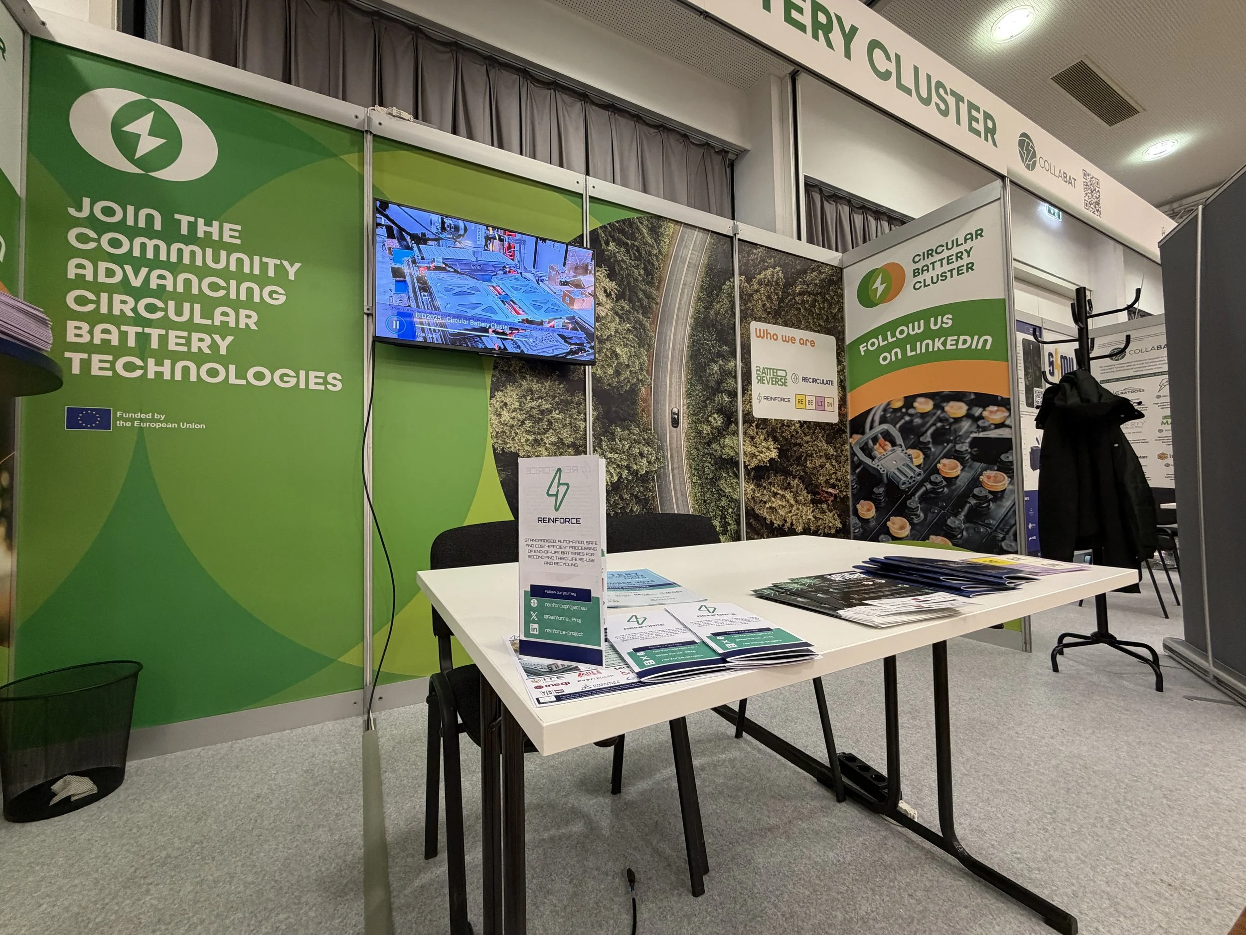 Trade show booth for Circular Battery Cluster featuring green banners with white text, a digital monitor displaying technical images, and a white table with brochures and flyers.