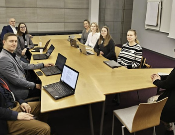 Group of nine people sitting around a conference table with laptops, in a modern office setting.