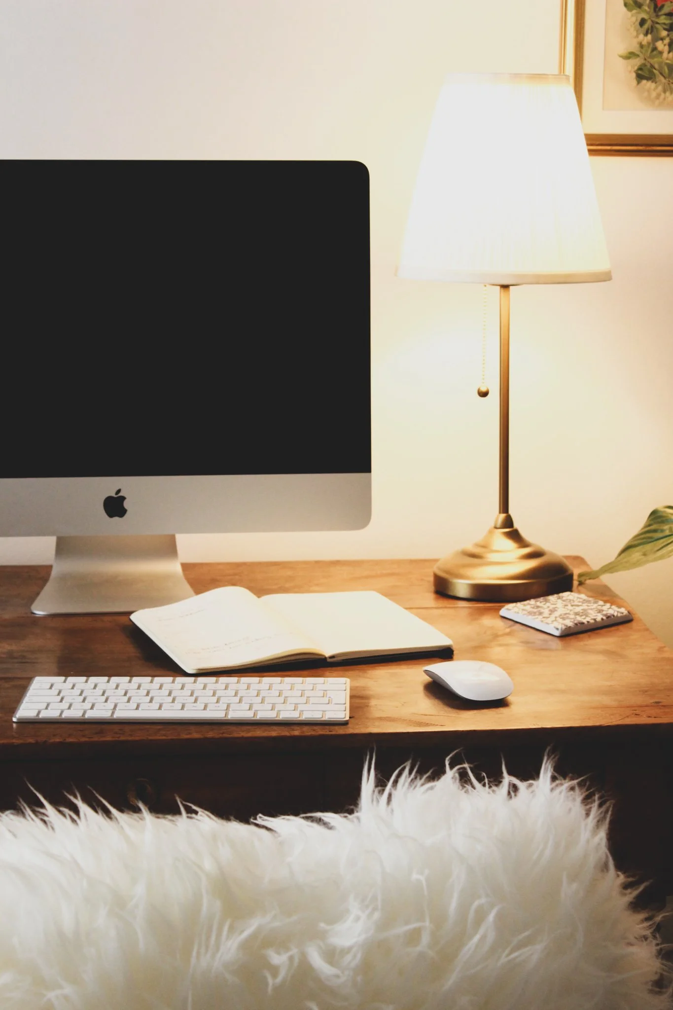 A desk setup with an iMac computer, an open notebook, a wireless keyboard, and a mouse. There is a cream-colored desk lamp turned on, a small notebook, and a potted plant on the right. The image has a fluffy white rug at the bottom.