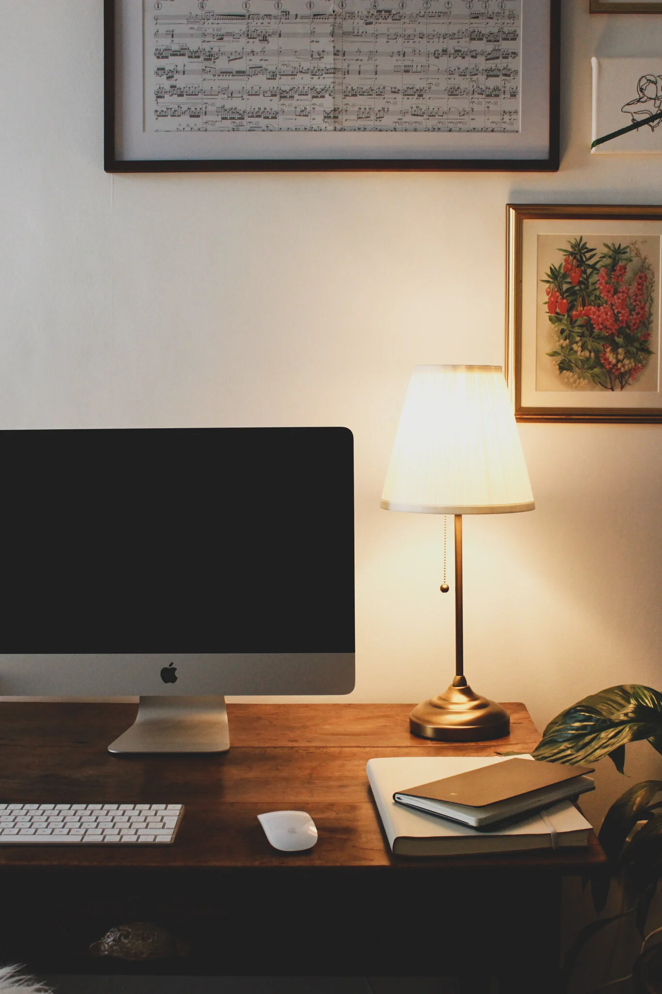 A home office desk with an iMac computer, a wireless keyboard, and mouse. A table lamp with a white shade is on the right side, and framed artwork, including a sheet music print and a botanical print, decorates the wall behind. There are also notebooks and a laptop on the desk, with a potted plant to the far right.