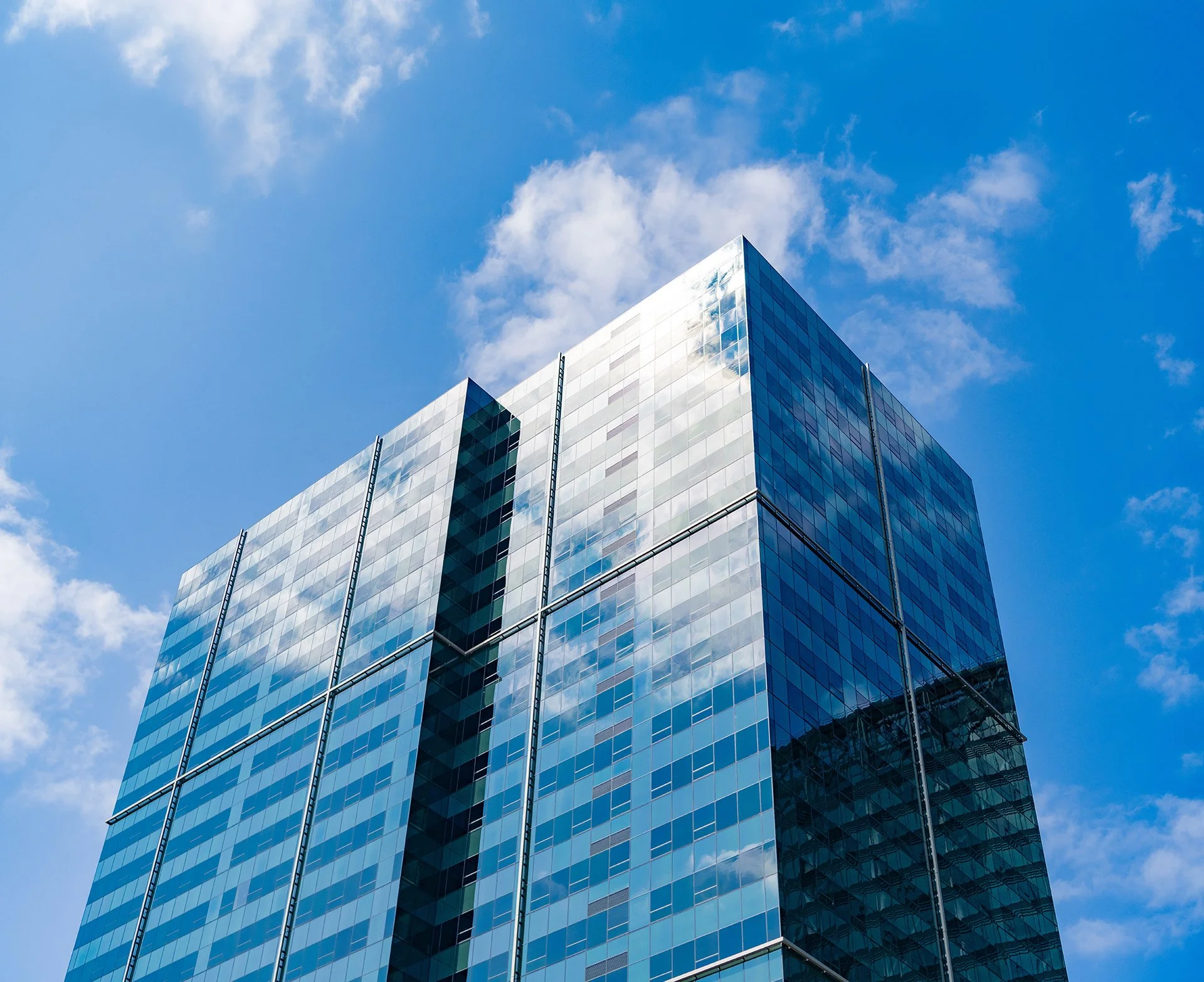A modern glass office building reflecting the sky and clouds, with a blue sky and scattered white clouds overhead.