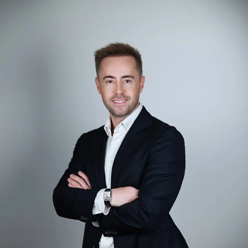 A young man with brown hair, wearing a white shirt and black blazer, standing with arms crossed and smiling against a plain, light-colored background.