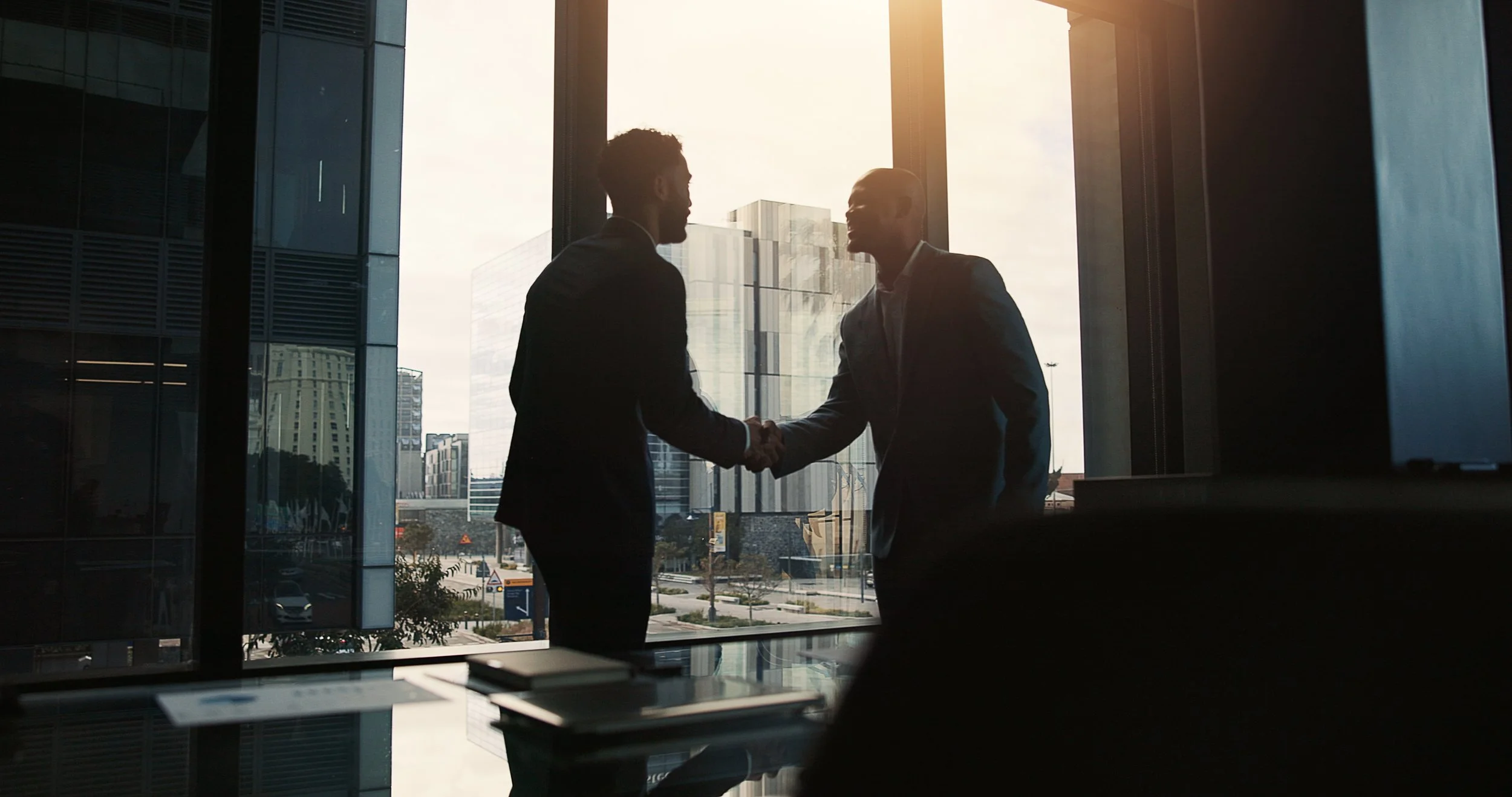Two businessmen shake hands in a high-rise office with large windows showing a city skyline at sunset.