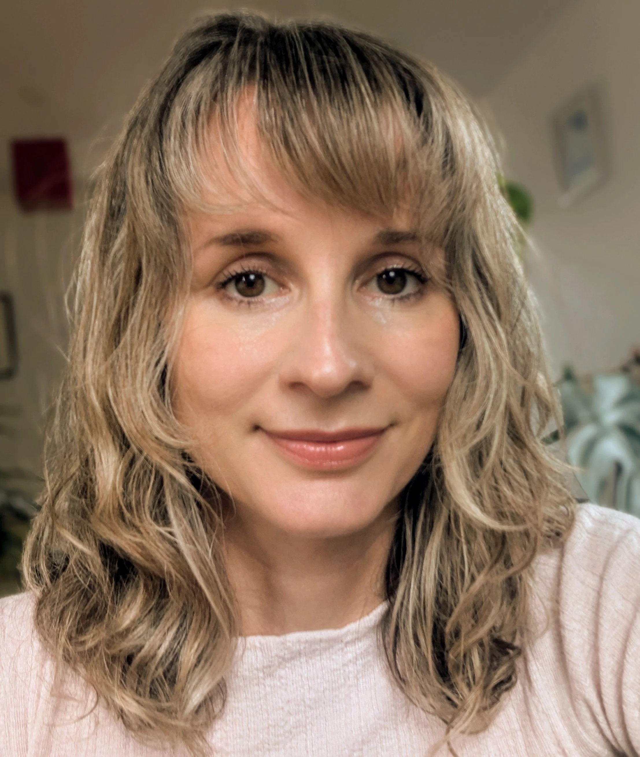 Close-up of a woman's face with light brown, wavy hair, wearing a light pink sweater, smiling softly.