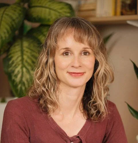 A woman with shoulder-length curly light brown hair, wearing a maroon top, sitting in a room with green houseplants and wooden shelves in the background.