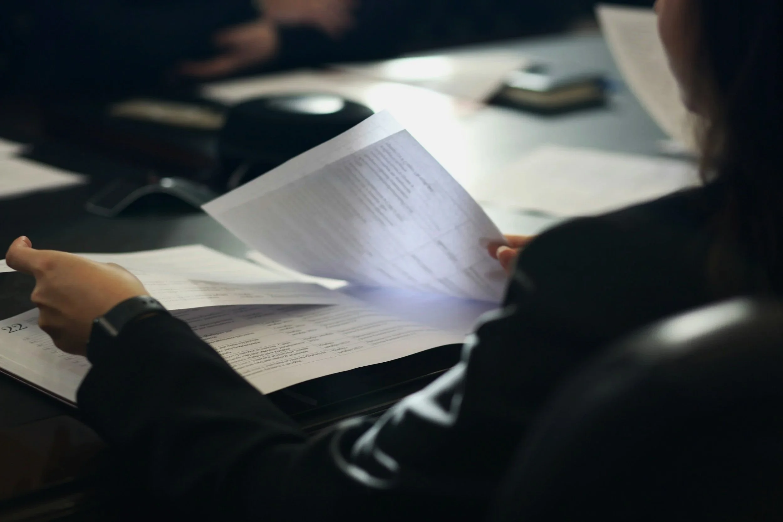 Person reading a printed document at a desk with other documents and office supplies around.