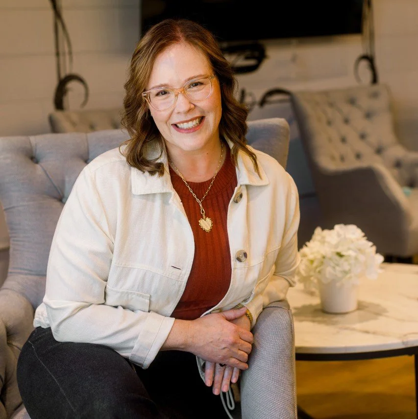 A woman with shoulder-length brown hair and glasses, smiling, wearing a white jacket over a rust-colored top, sitting in a comfortable armchair in a cozy room with a bouquet of white flowers on a table behind her.