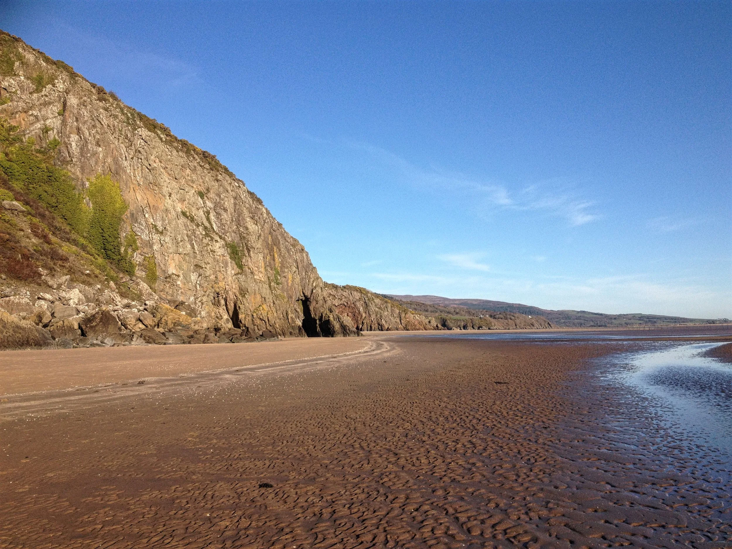 A sandy beach with tidal pools and rocks, backed by a steep, rocky hillside with sparse green vegetation, under a clear blue sky.
