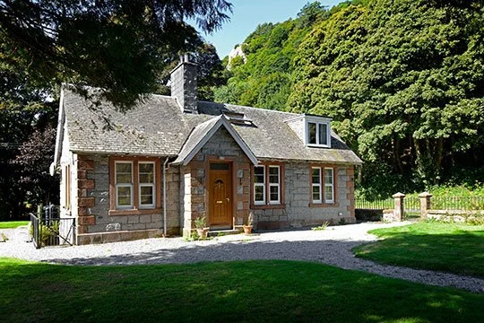 Stone house with brown door and multiple windows, surrounded by green trees and grass.