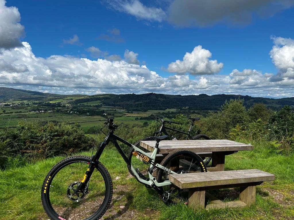 Two mountain bikes leaning against an outdoor wooden picnic table on a grassy hilltop with a scenic view of rolling green hills and a partly cloudy sky in the background.