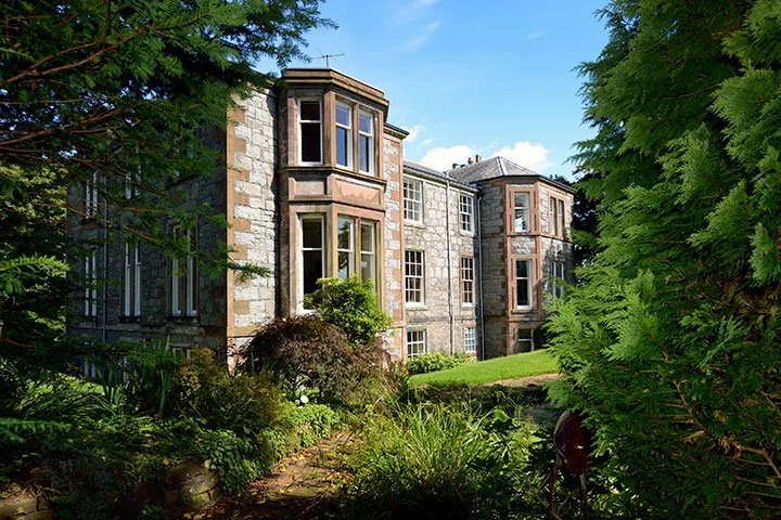 A large historic building with stone and brick exterior, multiple bay windows, surrounded by lush green trees and a landscaped garden.
