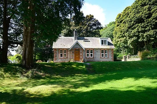 A stone house with a gray roof and front porch, surrounded by large trees and a lush green lawn.