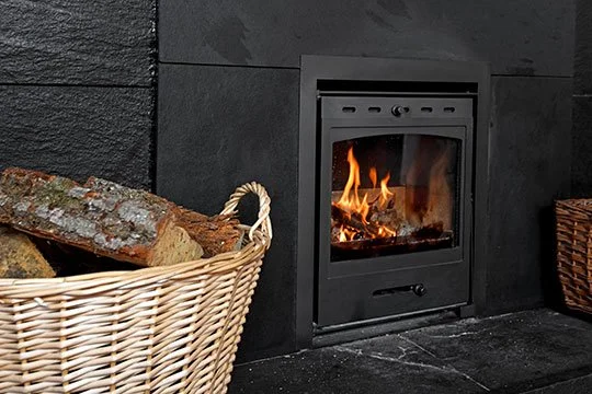 Indoor fireplace with a fire burning, surrounded by black wall paneling, with a basket of firewood to the left and a woven basket to the right.