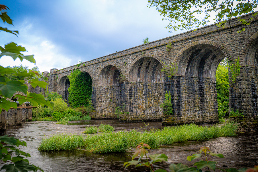 Stone bridge with multiple arches over a river, surrounded by greenery and trees, under a cloudy sky.