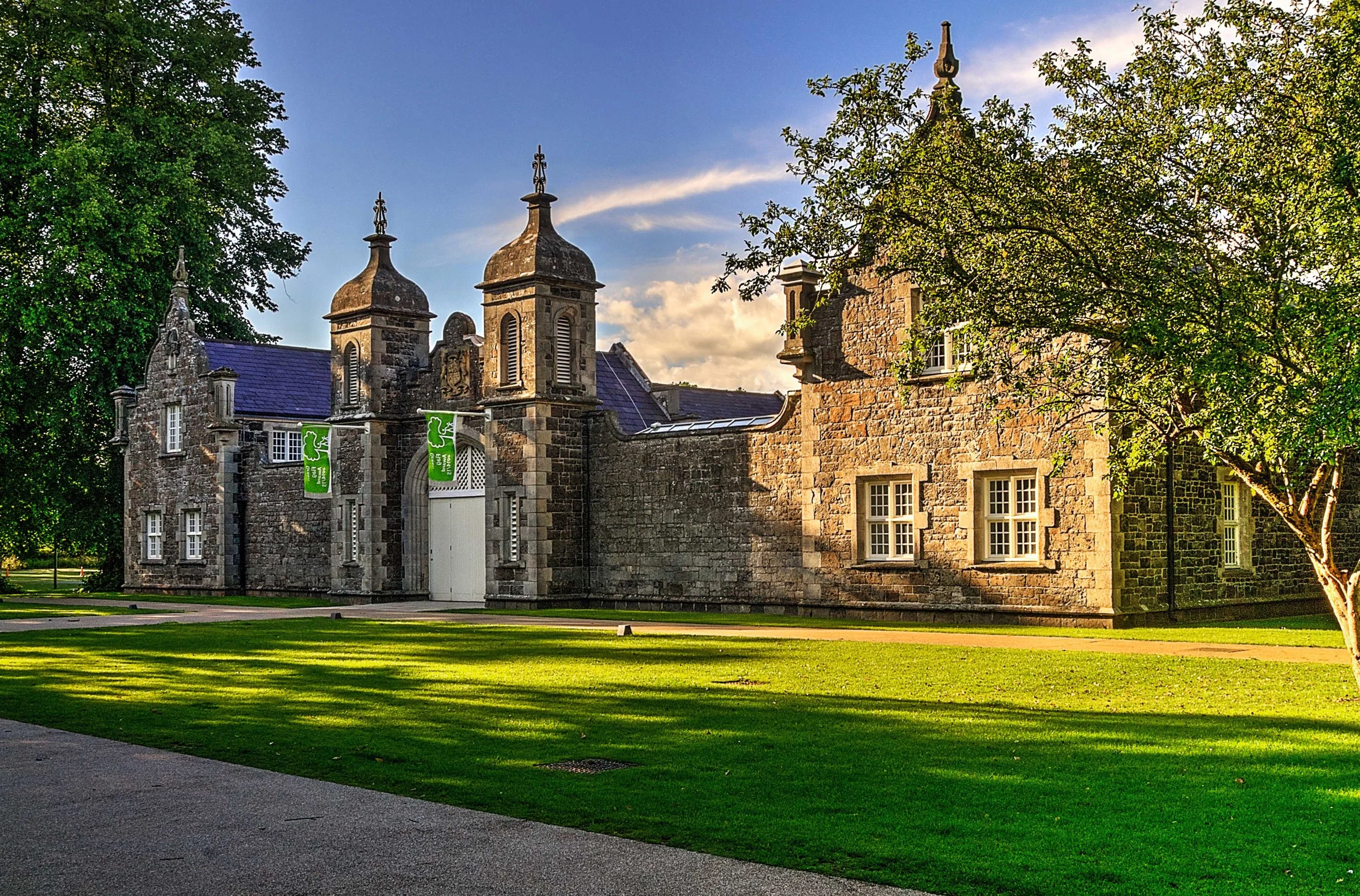 A historic stone castle with turrets and a large central door, surrounded by green trees and grass under a partly cloudy sky.