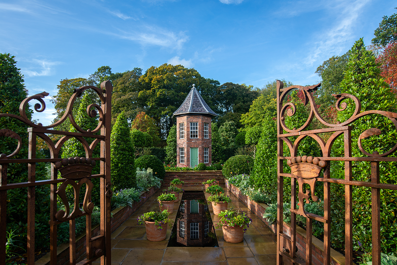 View of a garden with ornate metal gates, potted plants, and a brick tower with a pointed roof surrounded by lush green trees.