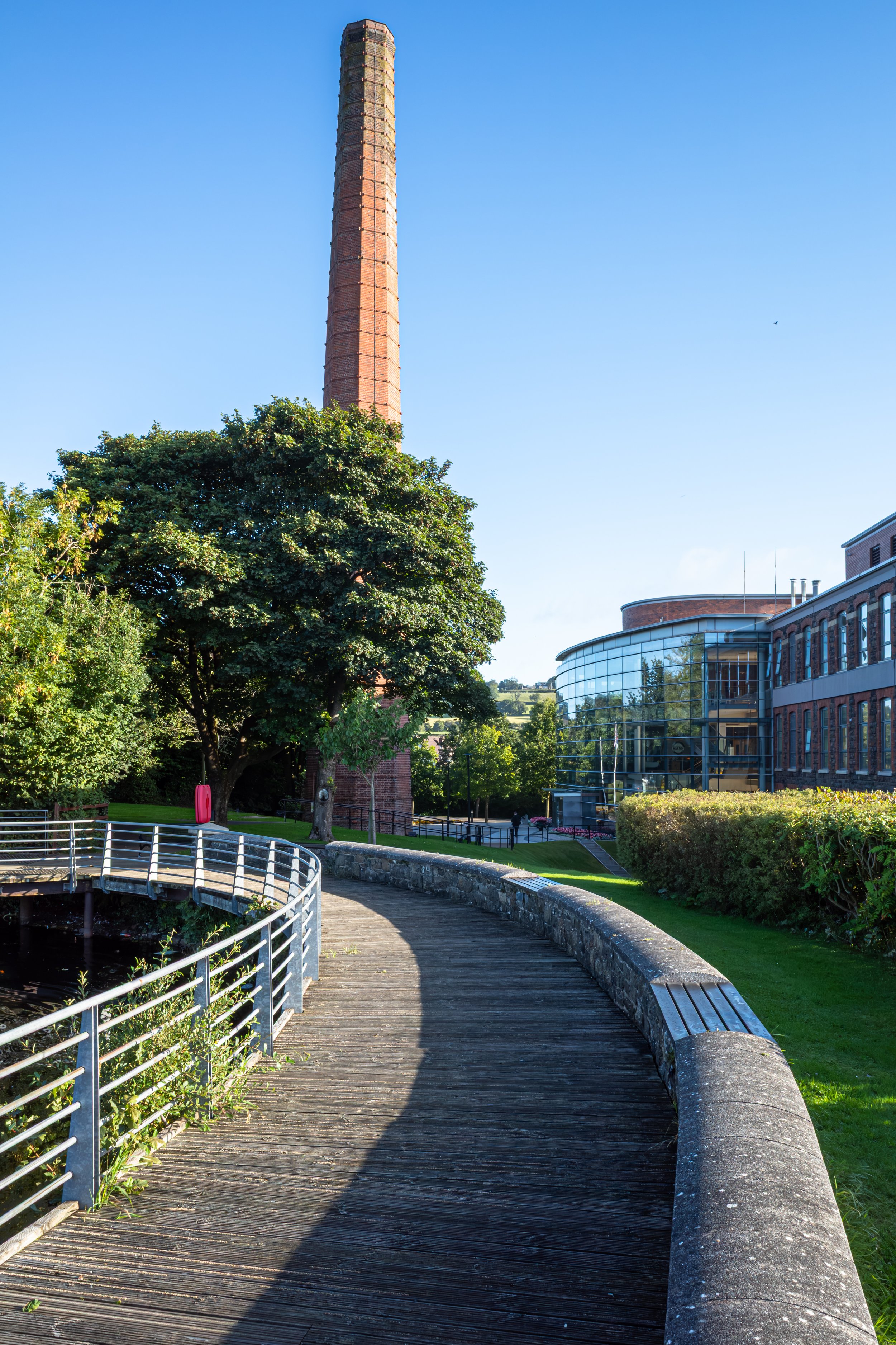 A scenic view of a city park with a wooden walkway, lush green trees, a river pond, and a tall brick chimney in the background against a clear blue sky.