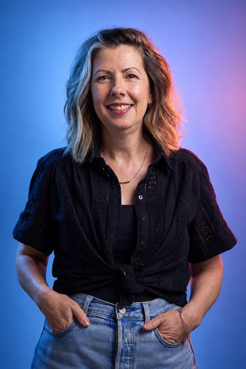 A woman with shoulder-length wavy hair smiling, wearing a black button-up shirt tied at the waist, and blue jeans, standing against a gradient background of blue and purple.